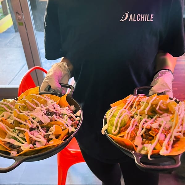 A person in an Al Chile Taco Bar shirt holds two skillets of loaded nachos with guacamole and crema.