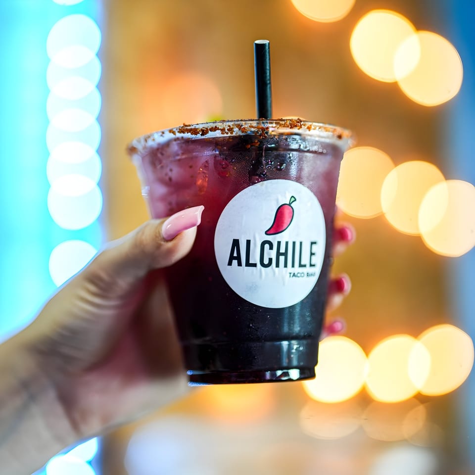 Hand holding an Al Chile Taco Bar drink with a chili-salted rim and black straw.