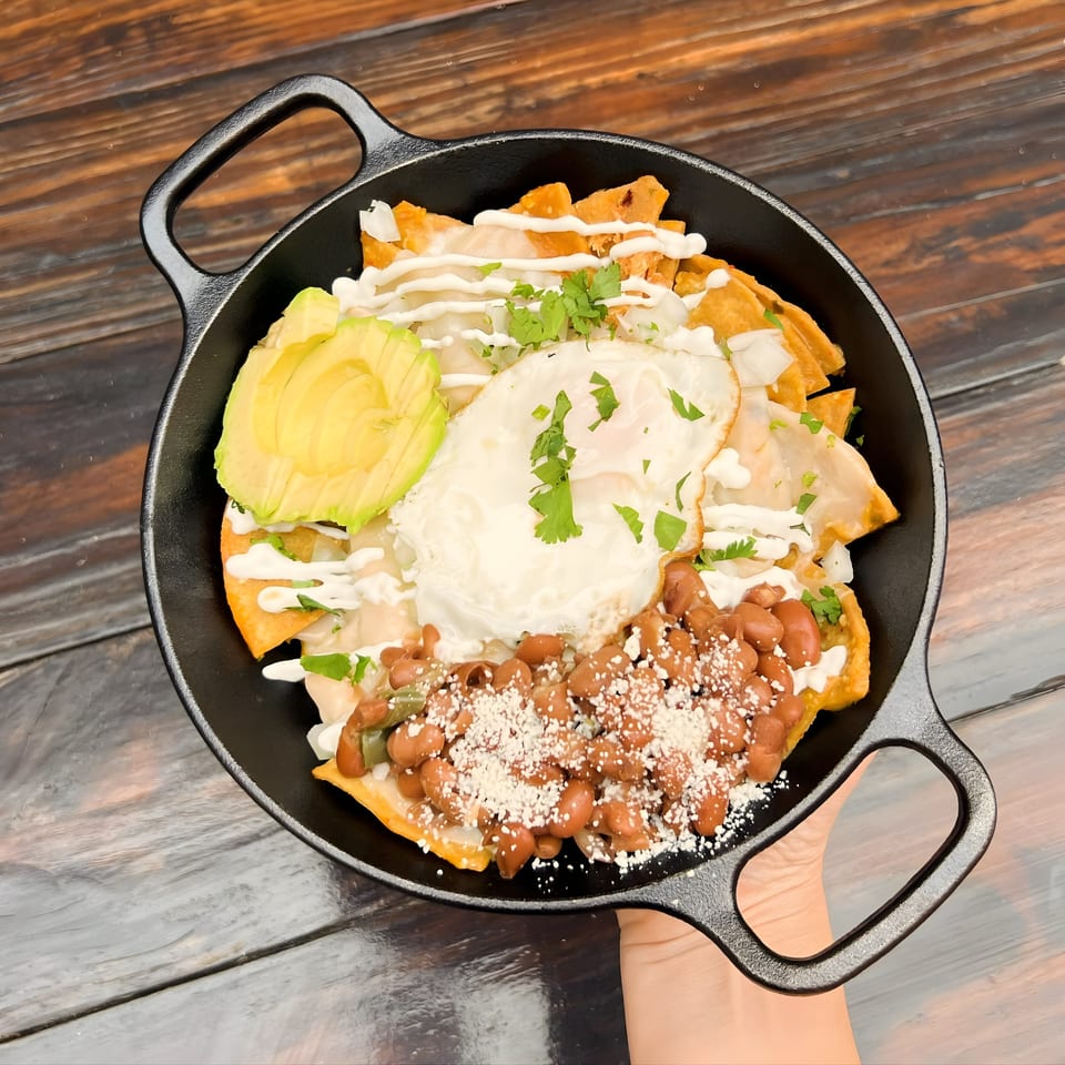 Chilaquiles with fried egg, avocado, beans, and crema in a black skillet on a wooden table.