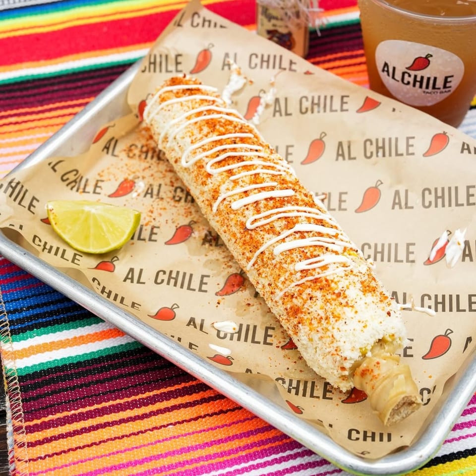 Elote with white sauce and chili powder, a lime wedge, and an Al Chile Taco Bar drink.
