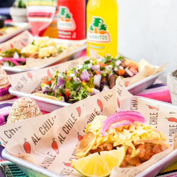 Al Chile Taco Bar's tacos with various toppings and Jarritos sodas on a colorful table.